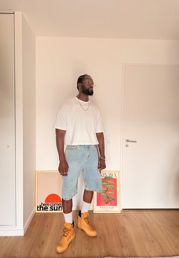 Homme portant un t-shirt blanc, un short en jean, des chaussettes blanches et des bottes jaunes, debout contre un mur avec deux affiches encadrées reposant sur le sol.