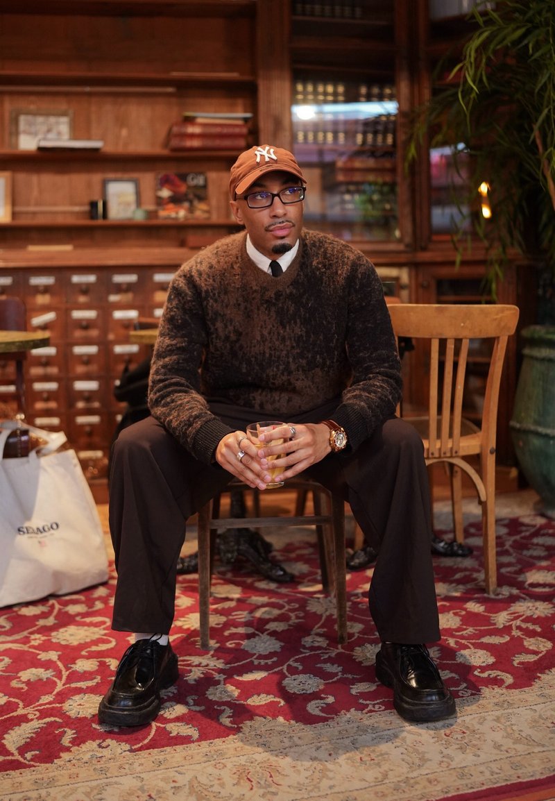 Homme portant des lunettes, un pull marron et une casquette, assis sur une chaise en bois, tenant un verre dans une pièce chaleureuse avec des étagères en bois et un tapis rouge à motif.