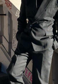 Costume noir à fines rayures avec une coupe ajustée. Le tissu a une légère brillance, avec une fermeture à un seul bouton et des revers crantés.