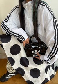 Černá mini kabelka z lakované kůže s bílým logem Adidas, bíl�á bunda s černými pruhy a černo-bílá puntíkovaná sukně na dřevěné podlaze.