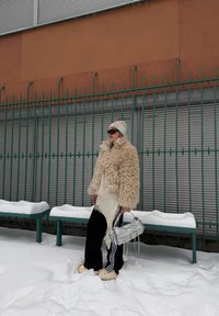 Personne portant une veste beige en fourrure, un bonnet crème, un pantalon noir et des lunettes de soleil, tenant un sac à main blanc, debout sur la neige près d'une clôture verte.