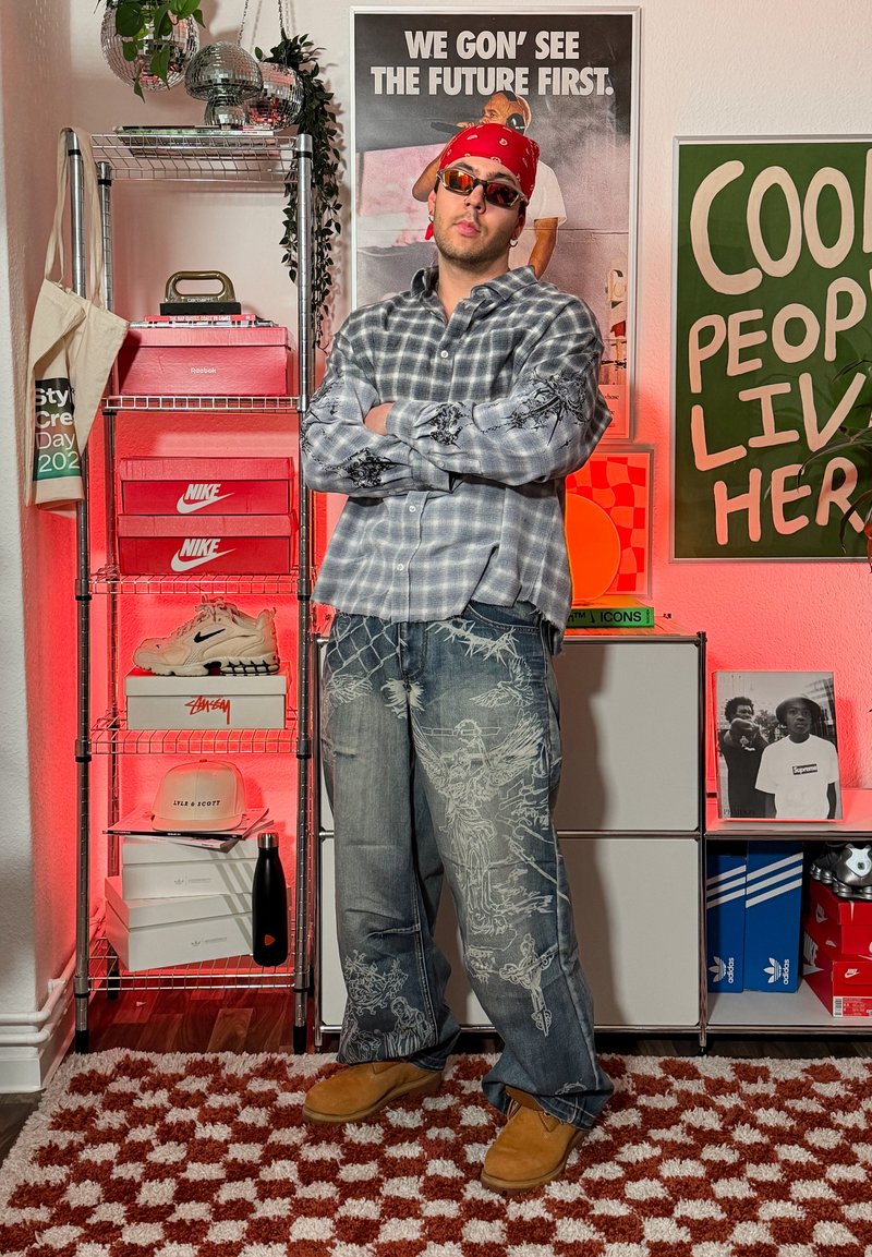Homme portant un bandana rouge, des lunettes de soleil, une chemise grise à motifs et un jean large, debout les bras croisés dans une pièce avec des baskets, des boîtes à chaussures et des affiches.
