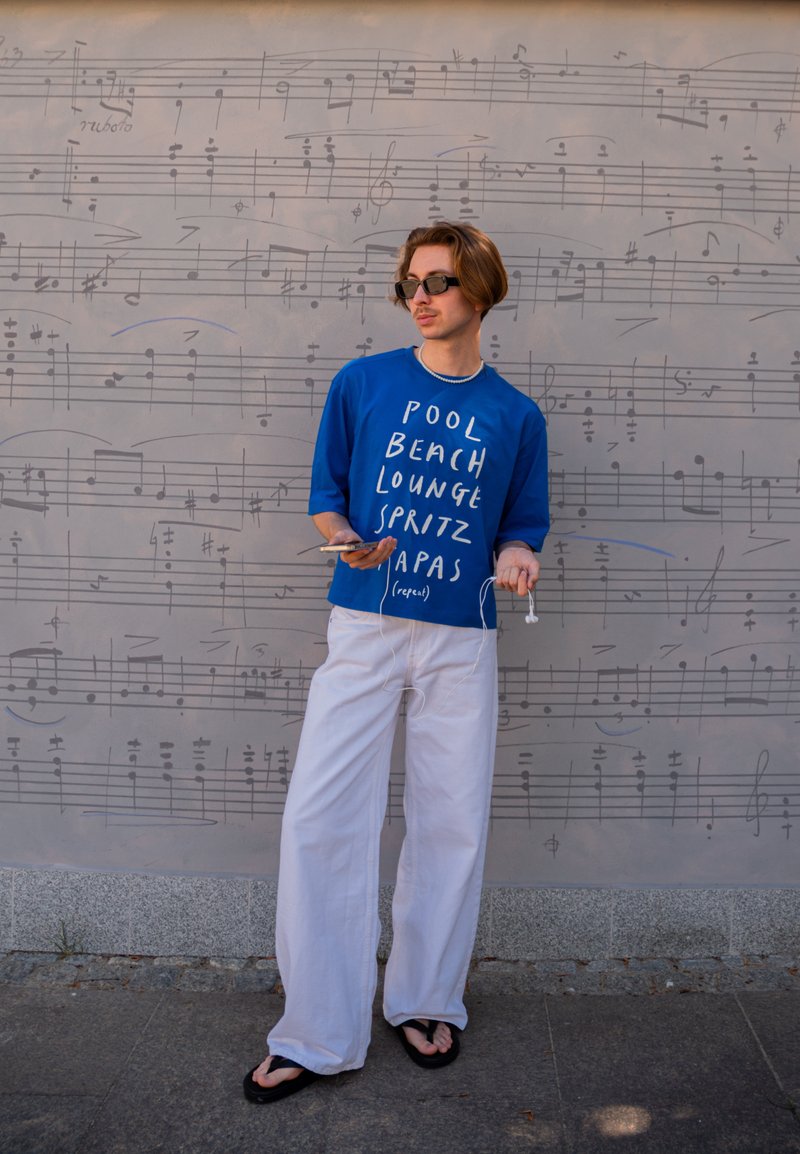 Jeune personne portant des lunettes de soleil, une chemise bleue avec du texte blanc, un pantalon blanc et des sandales noires, se tient près d'un mur couvert de notes de musique.