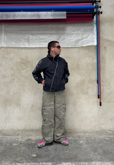 Chaqueta negra con cremallera, cuello alto y mangas estampadas, combinada con pantalones cargo sueltos de color gris claro y zapatos con acentos rosas.