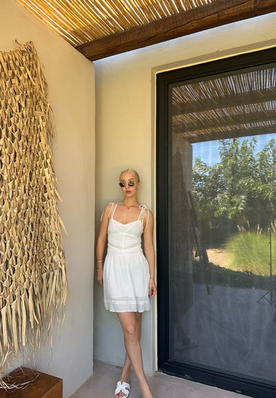 Vestido blanco de tirantes ajustables, con detalles de encaje y una falda fluida. Combinado con gafas de sol verdes redondas y sandalias blancas. Fondo natural.