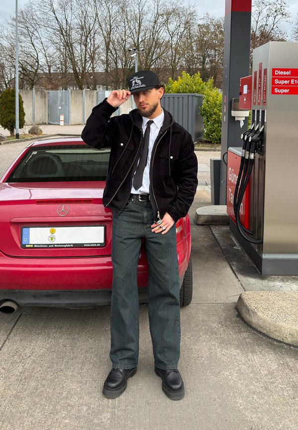 Uomo con giacca nera, cravatta e cappello accanto a una Mercedes rossa presso una stazione di servizio vicino ai distributori di benzina.