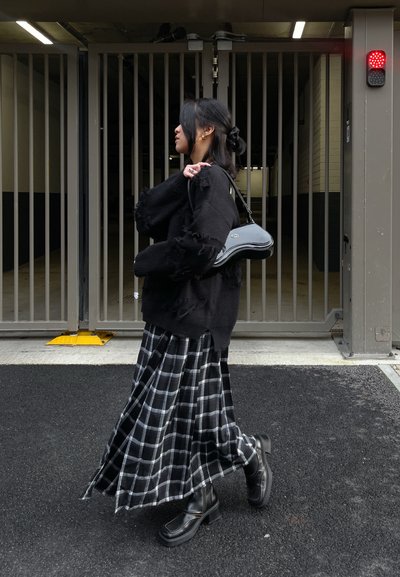 Suéter negro de gran tamaño con bordes deshilachados, colocado sobre una falda larga de cuadros en blanco y negro. Bolso negro de hombro y zapatos negros gruesos.