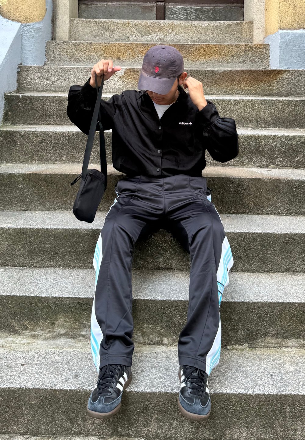 Svart skjorta med knappar, svarta och turkosa träningsbyxor, och svarta sneakers. Sitter på stentrappor med en liten svart väska.