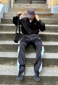 Svart skjorta med knappar, svarta och turkosa träningsbyxor, och svarta sneakers. Sitter på stentrappor med en liten svart väska.