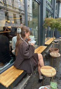 Une personne portant un grand blazer marron est assise sur un banc en bois, tenant une tasse à thé blanche. Près d'elle, il y a des tabourets en bois ronds et une boisson verte.
