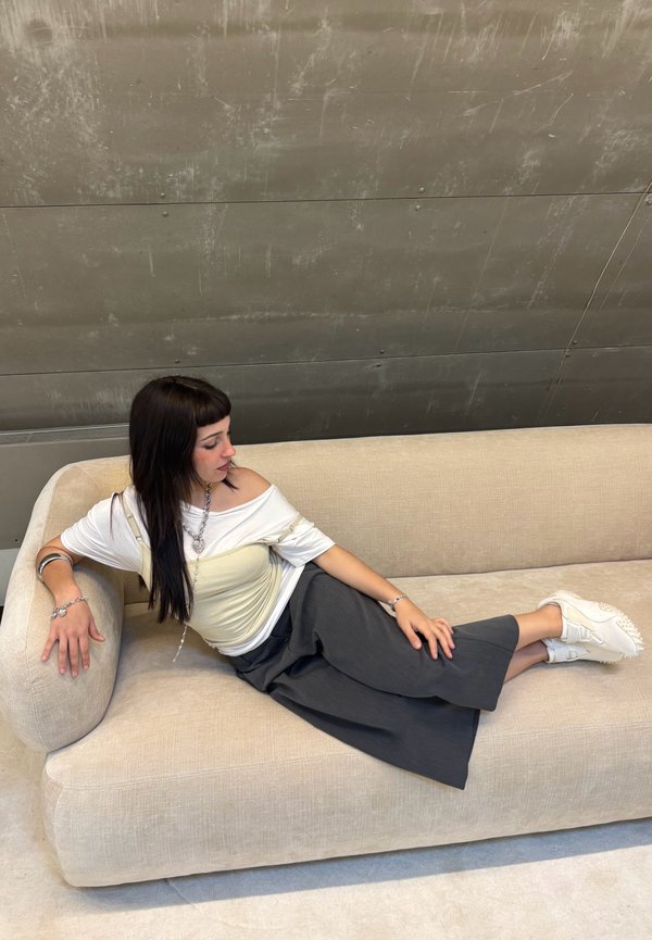 Ung kvinde med mørkt hår, der ligger tilbage på en beige sofa, iført hvid top, mørke vidde bukser, hvide sneakers og sølvsmykker, foran en grå væg.