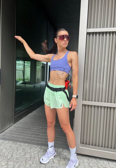 Conjunto atlético con un top corto púrpura, pantalones cortos verdes claros con detalles negros y calcetines blancos de Nike combinados con zapatillas. Los accesorios incluyen gafas de sol y un cinturón.