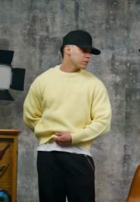 Pull côtelé jaune avec un col rond, superposé à une chemise blanche. Pantalon foncé avec un détail chaîne. Fond texturé gris.