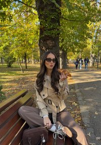 Béžový trenčkot s páskem, hnědé kalhoty a hnědé tenisky. Drží pe�čivo, sedí na dřevěné lavičce v parku s podzimním listím.