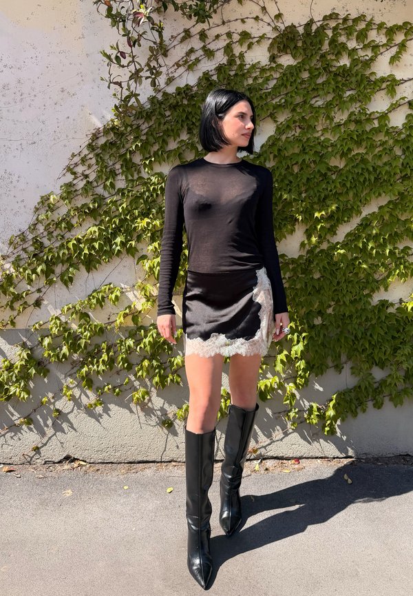 Frau mit schwarzem Bob-Haarschnitt trägt ein durchsichtiges schwarzes Langarmtop, einen schwarzen Rock mit Spitzenbesatz und kniehohe schwarze Stiefel und steht an einer mit Wein berankten Wand.