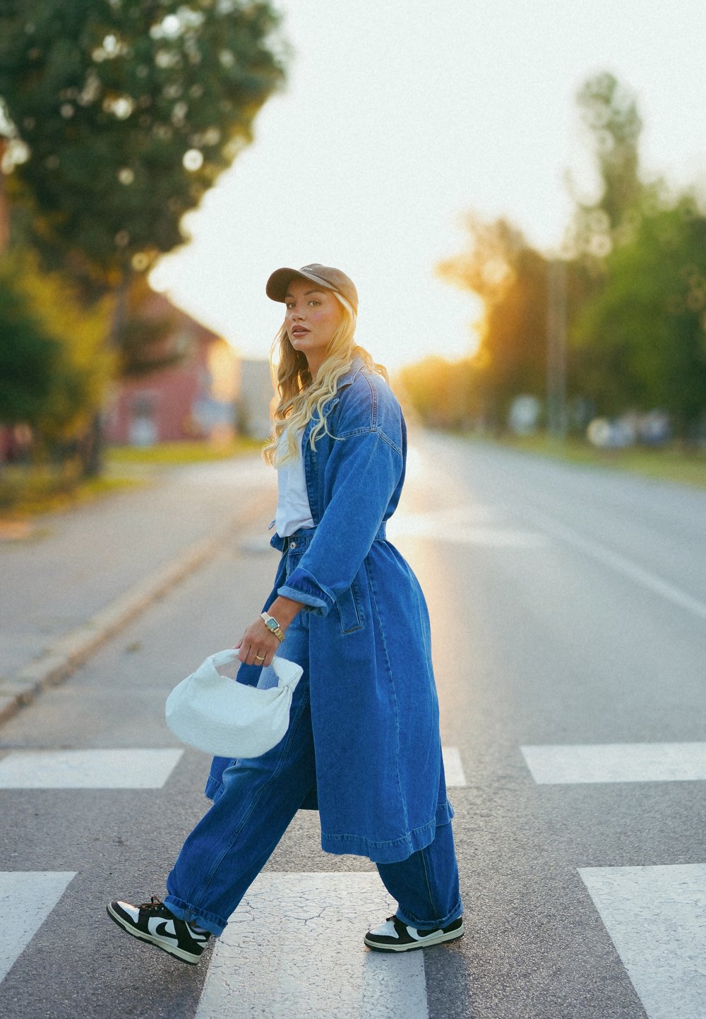 Casaco de ganga longo, calças de ganga largas a condizer, t-shirt branca e sapatilhas pretas. Segura uma bolsa de tecido branca. Passadeira ao fundo ao pôr do sol.