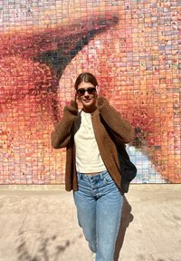 Cardigan de malha castanho sobre uma T-shirt branca, jeans azuis e óculos de sol escuros. O fundo apresenta um mosaico fotográfico colorido em tons quentes.