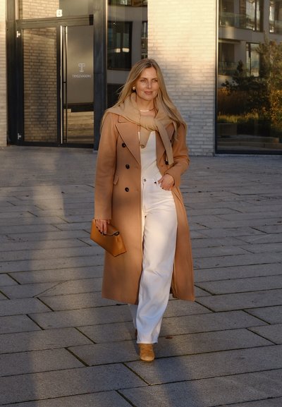 Abrigo largo beige de doble botonadura con botones oscuros, suéter crema, pantalones blancos, zapatos a juego y un pequeño bolso naranja.