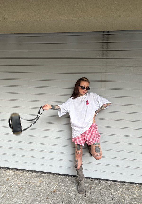 Jeune femme avec des tatouages porte des lunettes de soleil, un t-shirt blanc oversize, un short rouge à carreaux et des bottes grises, balançant un sac à main noir à son côté.