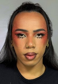 Retrato en primer plano de una persona con cabello largo y oscuro, maquillaje llamativo y piedras decorativas en el rostro. La piel tiene una textura suave.