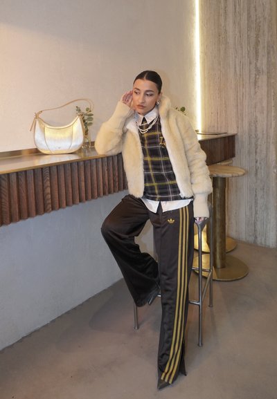 Mujer apoyada en el mostrador con chaqueta de piel crema, top a cuadros, collar de perlas y pantalones negros de Adidas con rayas doradas en un café moderno.