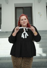 Μαύρη μπλούζα peplum με λευκή λεπτομέρεια με κορδόνι στο γιακά, σε συνδυασμό με παντελόνι λεοπάρ. Το μοντέλο έχει κόκκινα μαλλιά και κρατά το κορδόνι.