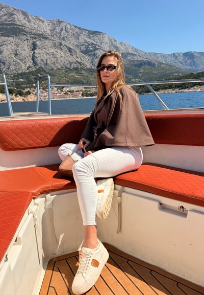 Joven mujer con gafas de sol, vestida con una chaqueta marrón y pantalones blancos, sentada en un banco rojo en un barco con montañas y mar de fondo.