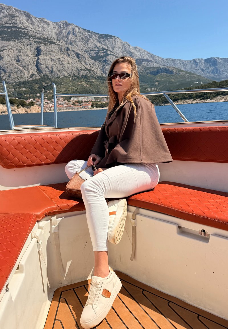 Νεαρή γυναίκα με γυαλιά ηλίου, που φοράει καφέ μπουφάν και λευκό παντελόνι, κάθεται σε ένα κόκκινο παγκάκι σε βάρκα με βουνά και θάλασσα στο φόντο.