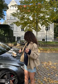 Bež kabanica preko tamnoplave veste, stojeći pored tamnosive automobila s dvije šalice kave na haubi, okružen jesenskim lišćem.