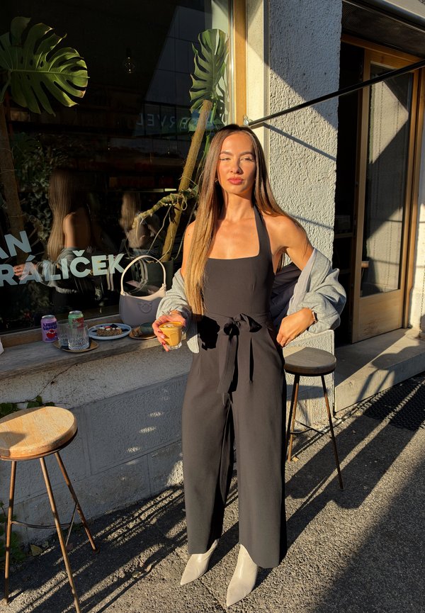 Una mujer con un mono negro y una chaqueta gris sostiene una bebida pequeña frente a una cafetería, bajo un sol radiante.