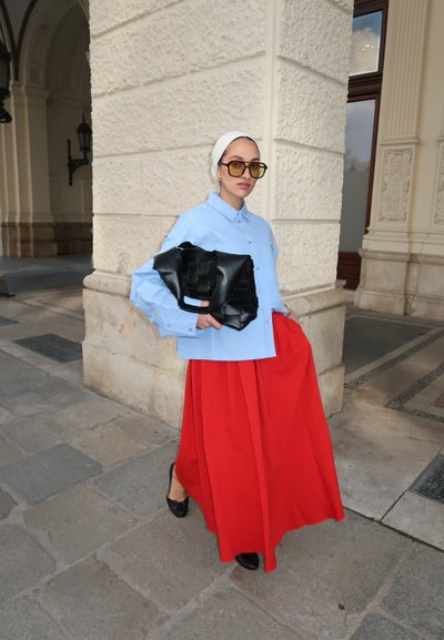 Camisa de botones azul claro, falda roja hasta el suelo y un bolso de mano negro de tejido. Gafas de sol grandes y cuadradas y zapatos planos negros completan el conjunto.
