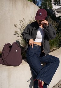 Casaco bomber cinza, top cropped branco, jeans de perna larga azul-escuro, saltos altos vermelhos, boné borgonha e uma bolsa redonda roxa no chão.