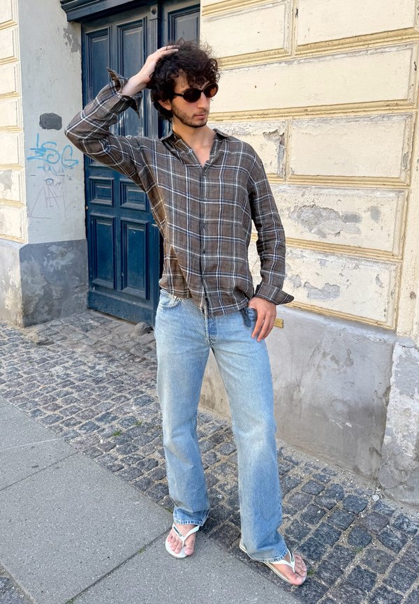 Man met zonnebril, gekleed in een bruin geruit overhemd, lichtblauwe jeans en witte slippers, staat op een kasseienstraat nabij een verweerde muur en een blauwe deur.