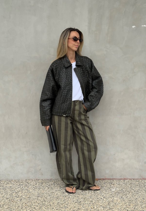 Femme portant une veste tissée noire, une chemise blanche, un pantalon large rayé olive et noir, des sandales noires, des lunettes de soleil, tenant une pochette noire près d’un mur gris.