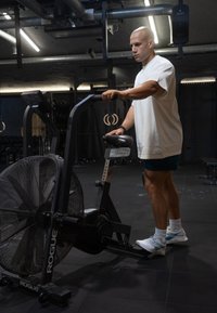 Luftcykel med en stor frontvifte, sort ramme, justerbart sæde og digitalt display. Bruger iført hvid skjorte og blå shorts.