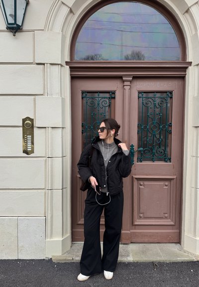 Chaqueta acolchada negra sobre un suéter gris, combinada con pantalones anchos negros y zapatillas blancas; de pie junto a una puerta marrón con detalles ornamentales.