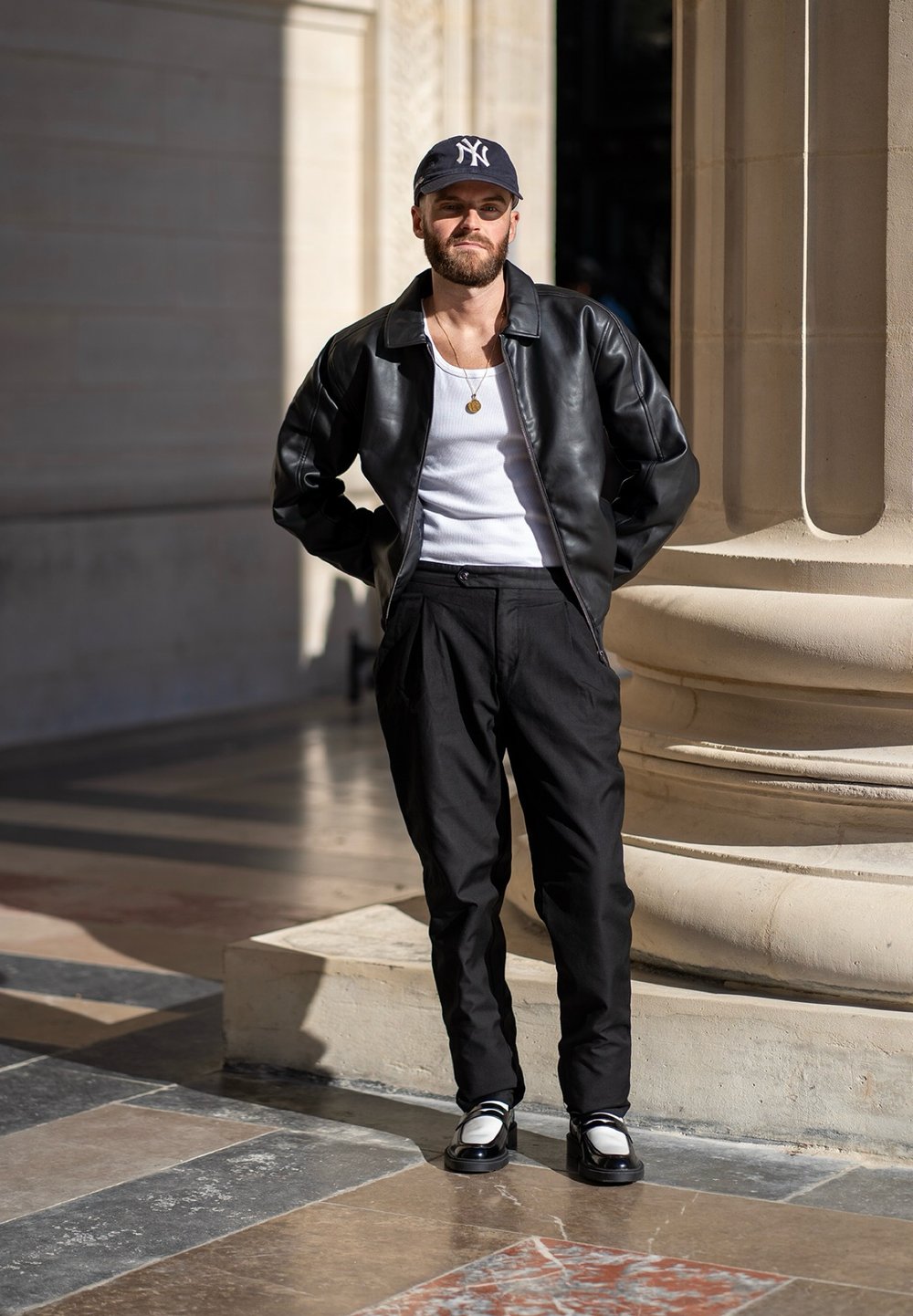 Μαύρο δερμάτινο τζάκετ πάνω από ένα λευκό λινό μπλουζάκι, συνδυασμένο με μαύρους παντελονιού ψηλής μέσης και δίχρωμα παπούτσια, στέκεται κοντά σε μια πέτρινη κολώνα.