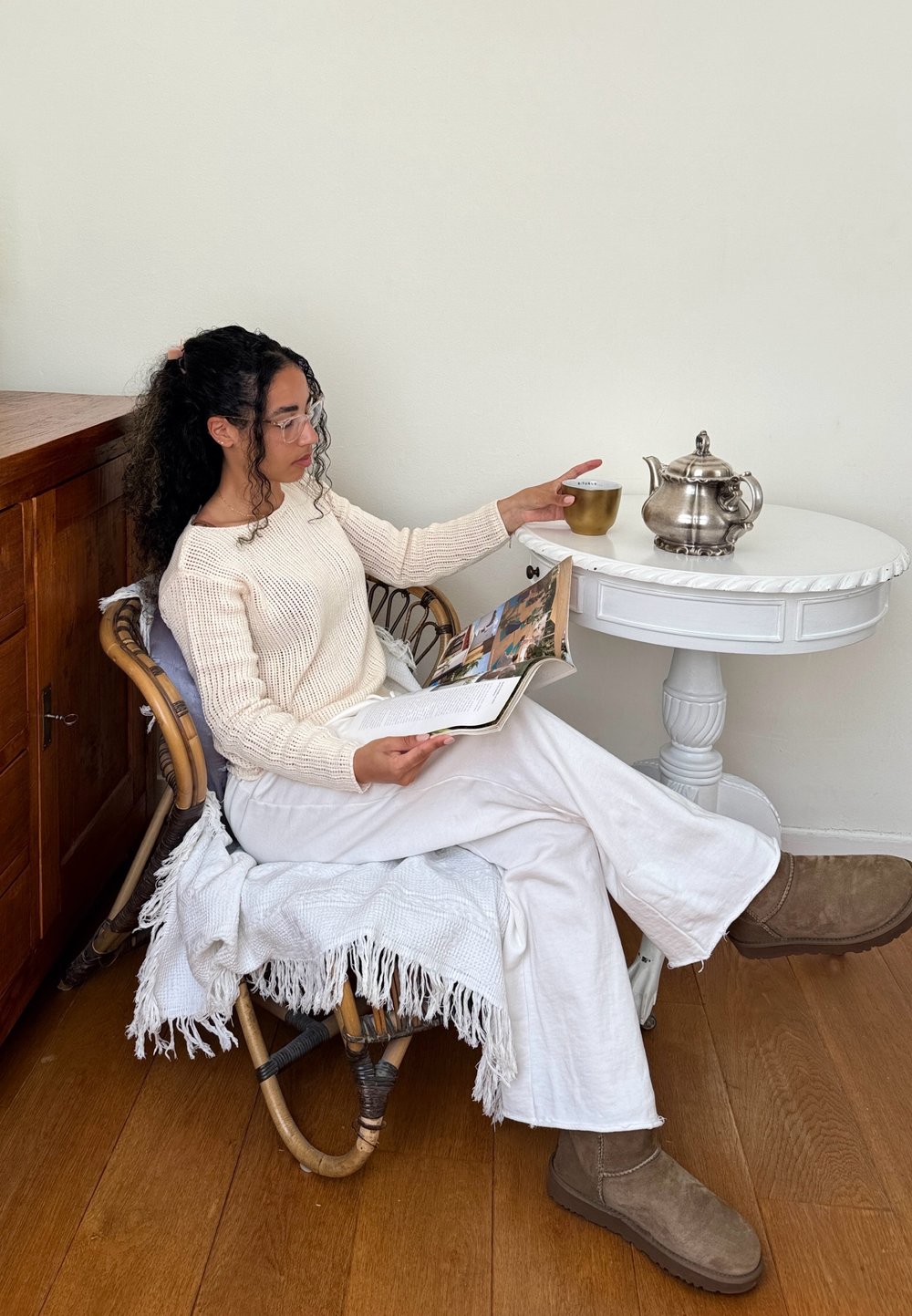 Sweater em malha creme, calças de linho brancas e botas de camurça castanhas. Sentada numa cadeira de rattan com uma manta branca com franjas, segurando uma revista ao lado de um conjunto de chá.