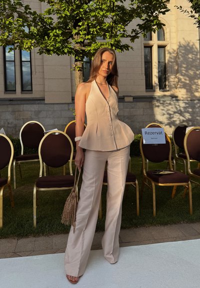 Top halter beis con botones y diseño peplum, combinado con pantalones acampanados color crema. Sosteniendo un bolso de flecos color marrón. Follaje verde y sillas al fondo.