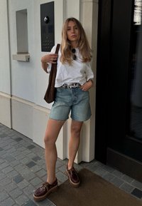 Γυναίκα στέκεται σε έναν τοίχο φορώντας ένα άνετο oversized λευκό T-shirt και ανοιχτό μπλε τζιν σορτσάκι με γυρισμένα τελειώματα, ενώ φοράει καφέ ψηλοτάκουνες σόλες.