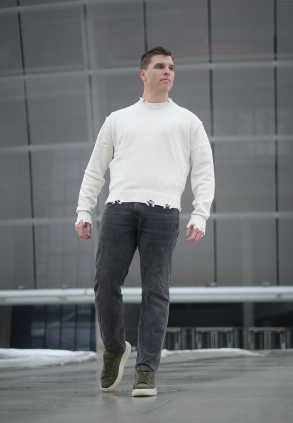 Mladi moški hodi na prostem in nosi bel izpran pulover, temne kavbojke in zelene superge v mestnem okolju.