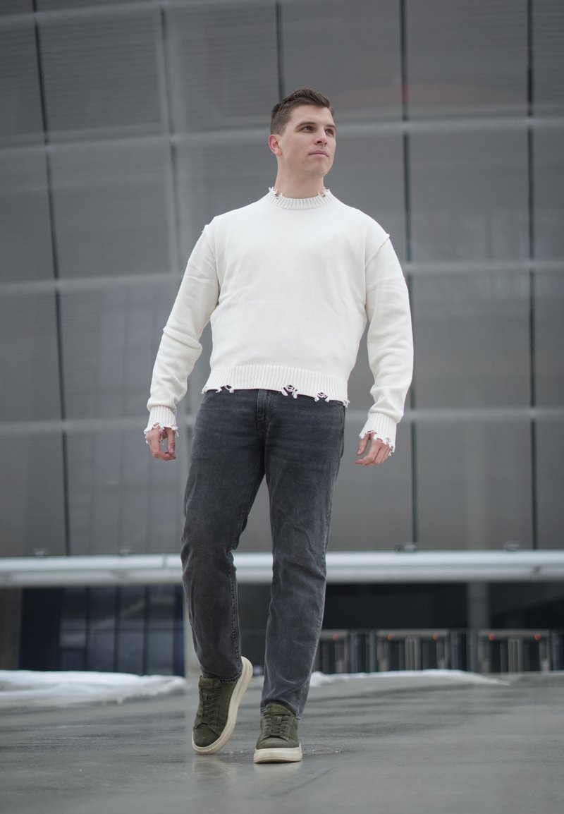Jeune homme marchant en plein air, portant un pull blanc usé, un jean foncé et des baskets vertes dans un cadre urbain.