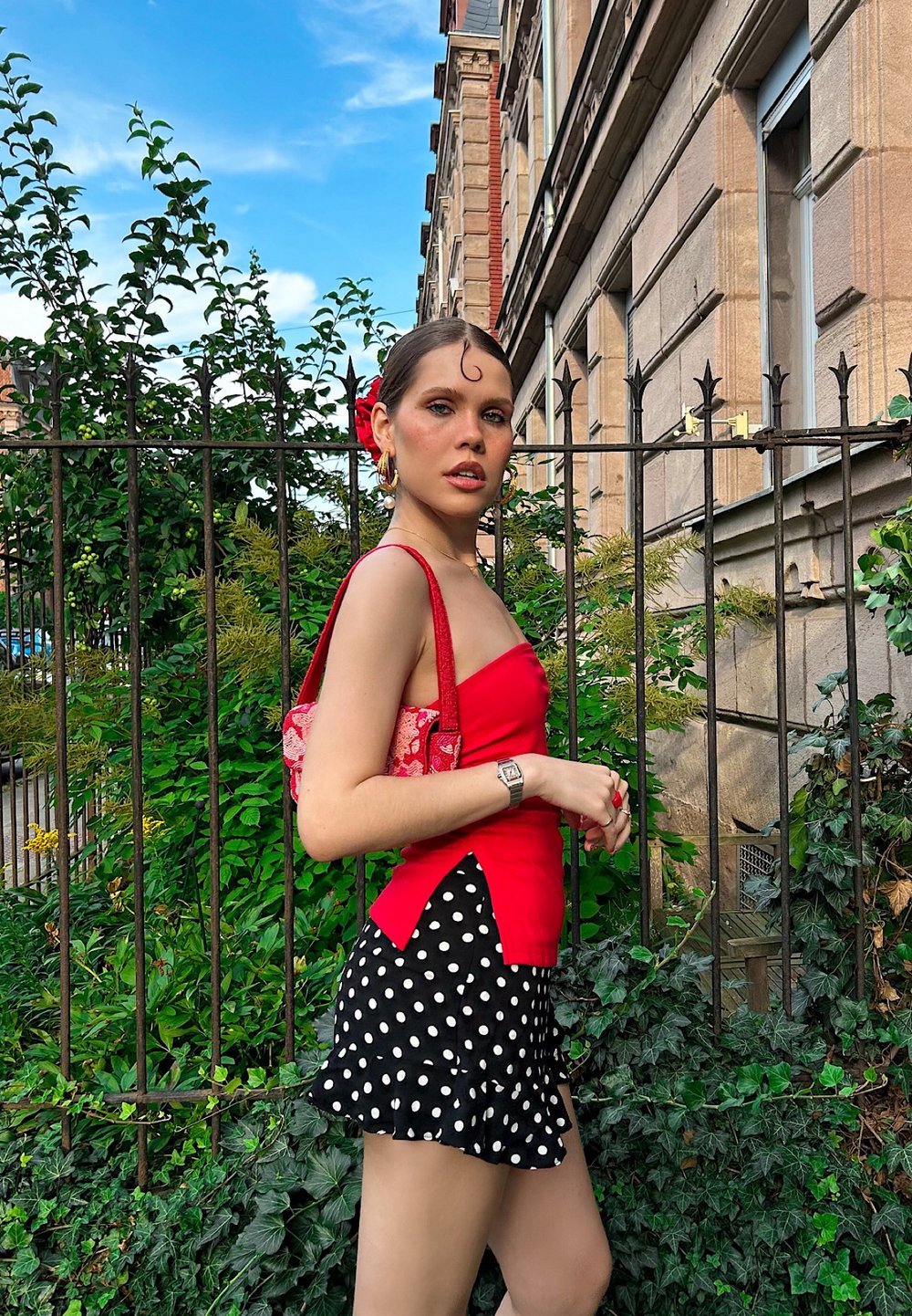 Rotes trägerloses Oberteil kombiniert mit einem schwarzen Rock mit weißen Punkten und einem gerafften Saum. Accessoires umfassen eine gemusterte Handtasche und eine Uhr.