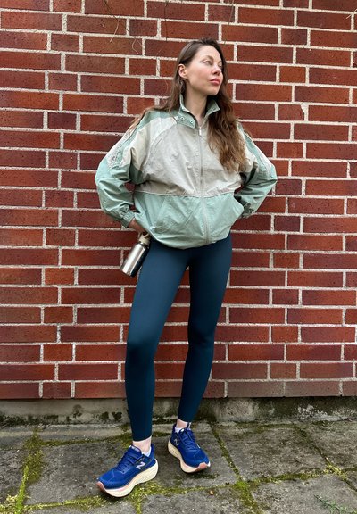 Chaqueta ligera en bloques de color verde y beige, combinada con leggings azul marino y zapatillas de correr azules, sosteniendo una botella de agua plateada.