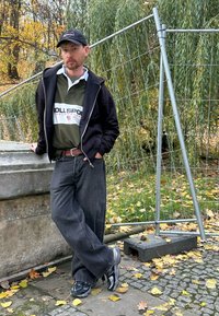 Czarna kurtka na zamek błyskawiczny noszona na zielonej koszulce polo z białym logo, ciemne dżinsy i czarno-szare trampki. W tle widać jesienne liście i ogrodzenie.