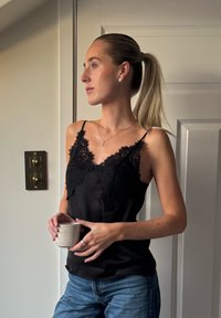 Camisola de cetim preta com detalhes em renda e alças finas. Modelo segura uma chávena de cor clara, usando jeans de ganga. Fundo neutro.