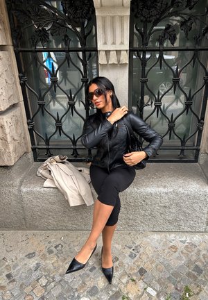 Chaqueta de cuero negra, leggings negros ajustados y tacones negros puntiagudos. Abrigo beige claro doblado cerca. Fondo de forja intrincada.