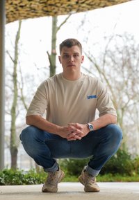 Beige t-shirt med kort ärm och texten "SPRING" i blått, i kombination med blå jeans och beige sneakers, modell som hukar i en utomhusmiljö.