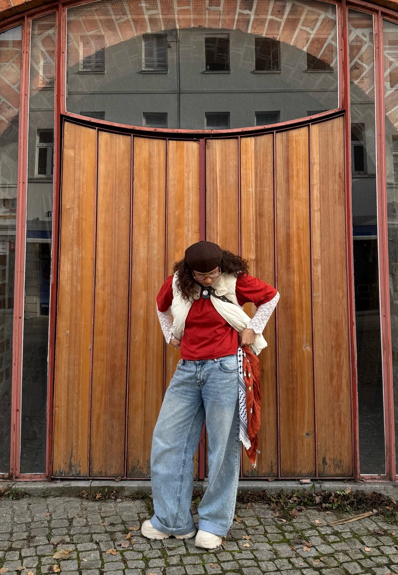 Une personne se tient devant une porte en bois, portant une chemise rouge, un gilet blanc et un jean à jambes larges, accessoirisé d'une écharpe à motifs.