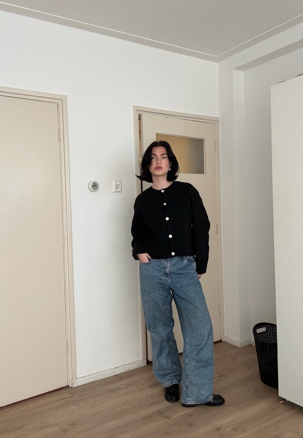 Femme aux cheveux noirs et courts debout à l'intérieur, portant un pull noir boutonné, un jean bleu à jambes larges et des chaussures noires, les mains dans les poches.